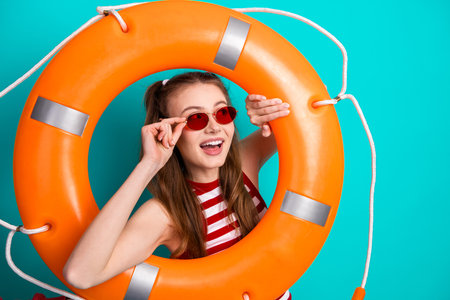 Cheerful young woman posing with an orange life preserver on a vibrant teal background showcasing...