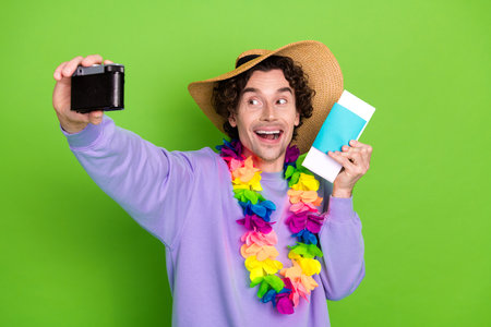 Happy young man with a lei and hat taking a selfie on a green background while holding a travel...