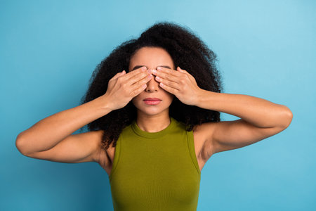 Attractive young woman in casual green top covering eyes with hands while posing against vibrant blue backgroundの写真素材