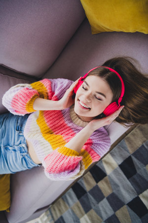 Charming woman relaxing on sofa wearing colorful sweater and listening to music with headphones,...
