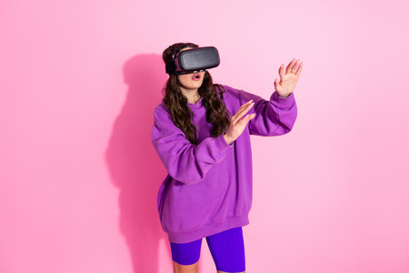 Young woman experiencing virtual reality wearing stylish purple clothing against pink backdrop,...