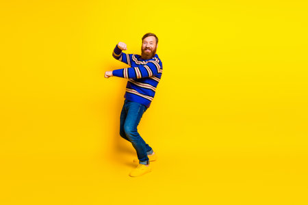 Cheerful young man with a red beard dancing playfully in a colorful outfit against a bright yellow backgroundの写真素材