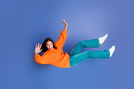 Young woman in orange sweater falling with surprised expression against blue background displaying modern casual styleの写真素材