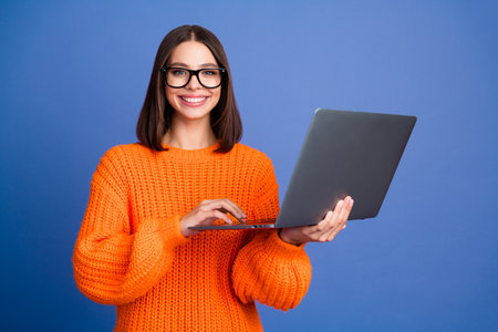 Young woman in orange sweater using laptop against blue background, smiling confidently and enjoying...