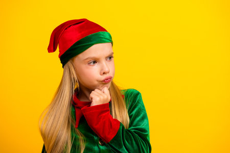 Young girl in festive elf costume on yellow background, pondering expression, capturing Christmas...