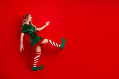 Festive young woman dressed as a Christmas elf posing against red background, spreading cheer with a...