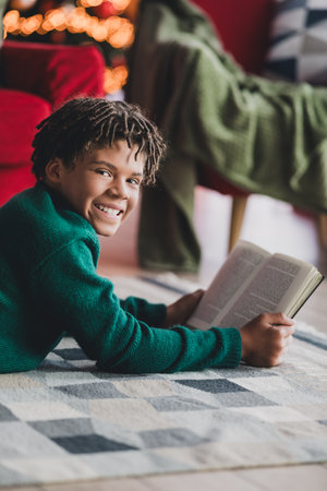 Cheerful boy reading a book on a cozy rug in a festively decorated room with warm Christmas lights...