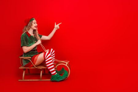 Playful girl in elf costume pointing to side while sitting on a vintage wooden sleigh against bright red backgroundの写真素材