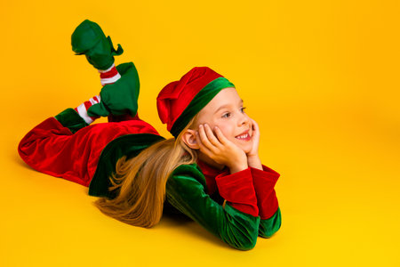Playful girl in festive green and red elf costume smiling cheerfully against bright yellow background during holiday seasonの写真素材
