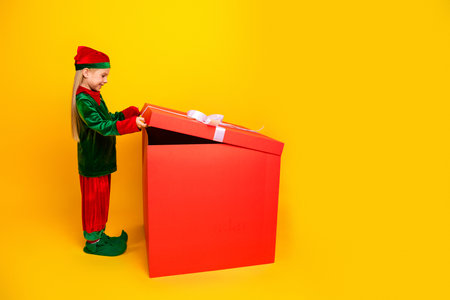 Smiling child dressed as a festive elf opening a giant red gift box against a bright yellow...