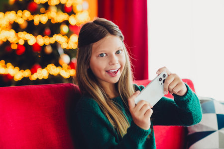 Cheerful girl plays on her phone beside a glowing Christmas tree at home with festive lights and...
