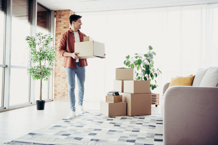 Young man moving into a modern apartment carrying cardboard boxes in a bright living room full of...