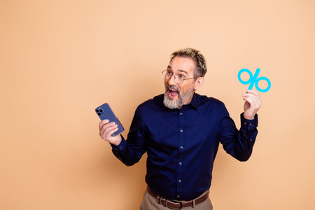 Middle-aged businessman promoting a sale with a percentage symbol and smartphone against a stylish beige backgroundの写真素材