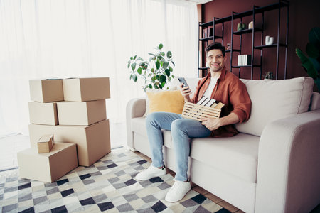 Young man happily sitting on a couch in a modern apartment surrounded by moving boxes, enjoying a...