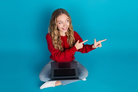 Adorable young girl in a red sweater enjoying leisure time with laptop on blue background