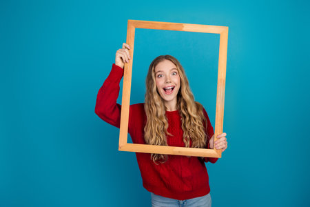 Adorable blonde girl holding a wooden frame, wearing a stylish red sweater against a vibrant blue...