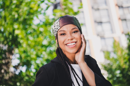 Bright smiling woman in stylish headwrap and blazer enjoys sunny city park outing showcasing modern...