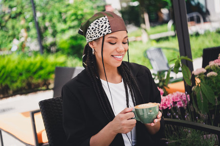 Playful stylish woman in a headscarf enjoys a coffee on a sunny outdoor cafe moment surrounded by greenery and blooms a chic casual scene of modern city lifeの写真素材