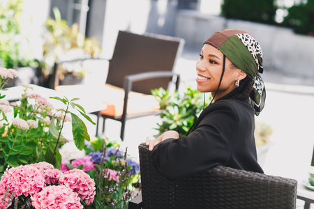 woman in headscarf smiles at an outdoor cafe garden filled with pink flowers enjoying a bright sunny...