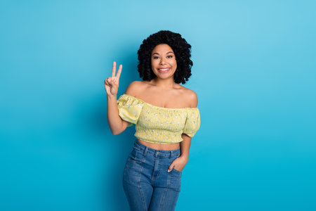 Joyful young woman with curly hair making a peace gesture, outdoors casual summer look on blue...