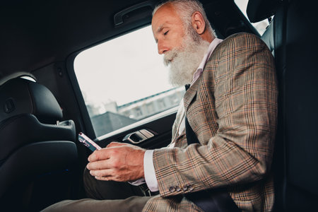 Mature Businessman in a Stylish Suit Utilizing a Smartphone Inside a Car in an Urban Environment...