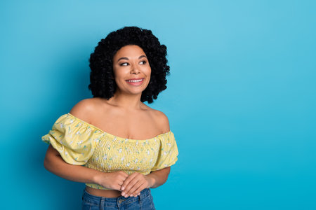Pretty woman with curly hair and yellow top smiling against blue background showing cheerful and...