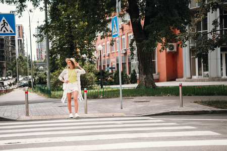 Young woman with sunhat stands at crosswalk in sunny urban setting enjoying a fashionable summer day shopping walking and travelingの写真素材