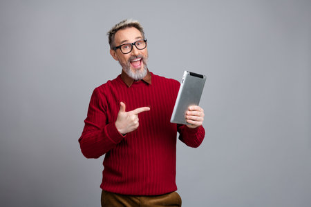 Enthusiastic mature businessman promoting technology with vibrant gesture in sweater holding tablet...