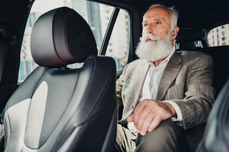 Senior businessman with beard sitting in cars backseat on urban street, dressed elegantly in jacket