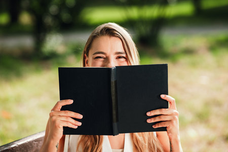 Young woman enjoys a sunny outdoor moment reading a black notebook while sitting on a park bench...