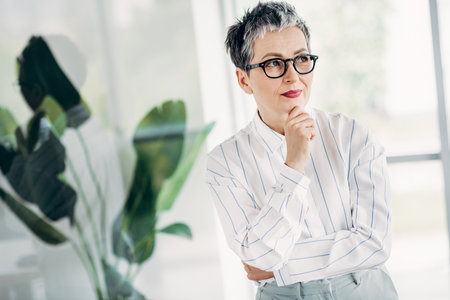 Confident Mature Woman Posing Thoughtfully in a Stylish Office Space with Formal Attire Reflecting...