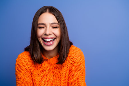 Laughing young woman in vibrant orange sweater posing against purple background for fashion and lifestyle themesの写真素材