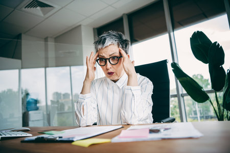 Qualified businesswoman in stylish office setting appearing stressed, seated at desk with papers and...