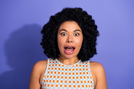 Excited young woman with curly black hair expressing surprise against a purple background in casual...