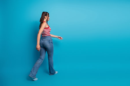 Young woman in denim jeans walking confidently with a playful demeanor against a bright blue...