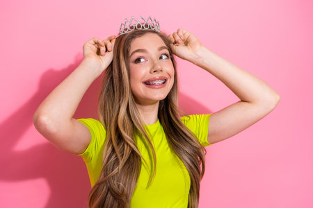 Lovely young girl with crown posing confidently on pink background, radiating cheerful beauty and youthful charmの写真素材