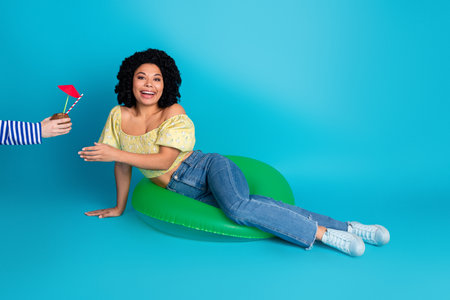 Charming young woman on a green inflatable cushion in casual attire against a vibrant blue...