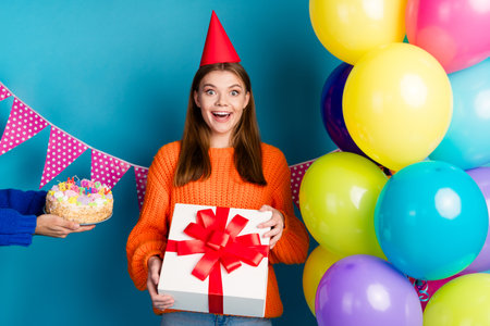 Charming young woman in orange sweater posing with gift and balloons against vibrant blue background