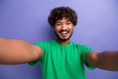 Cheerful young man in a green shirt taking a selfie against a purple background exuding positivity and energyの写真素材