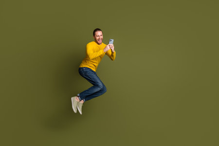 Young man in casual yellow sweater holding smartphone jumping joyfully against minimalist green...