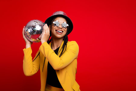 Woman holding a disco ball wearing stylish attire against a vibrant red background, exuding joy and...