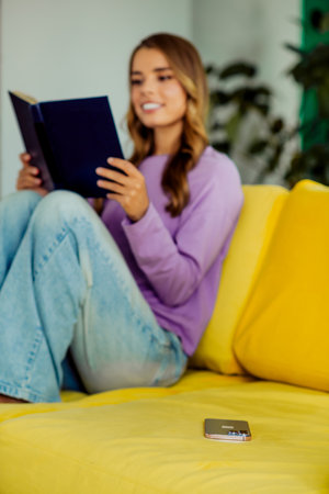 Young woman enjoying a book on a bright yellow sofa within a cozy living room space, embracing...