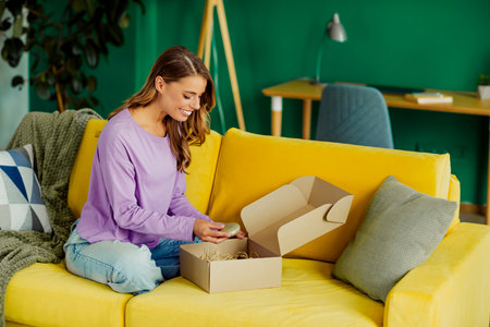Smiling young woman unpacking a parcel while sitting on a cozy couch in a vibrant living room