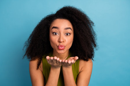 Stylish young woman blowing a kiss against a blue background, expressing happiness in a trendy green top and natural curlsの写真素材