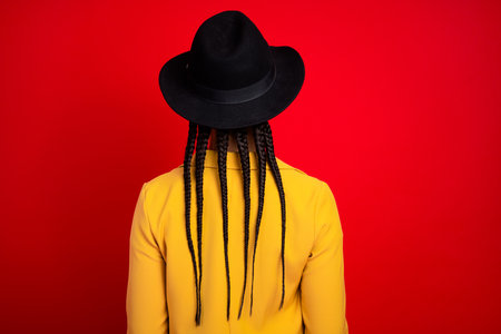 Back view of a stylish woman with braided hair wearing a hat and yellow jacket against a vibrant red...