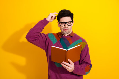 Young man in colorful sweater and eyeglasses reading a book with a thoughtful expression, set...