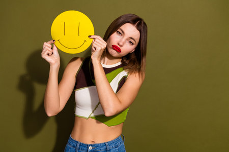 Young woman holding a yellow smiley face sign while showcasing contrasting emotions against a khaki backgroundの写真素材