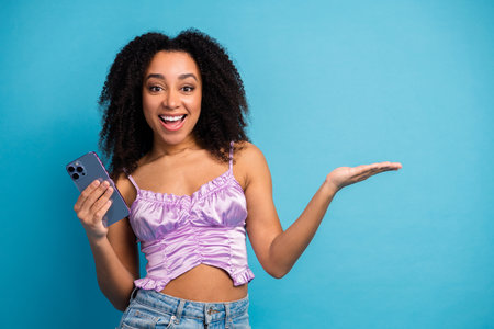 Young stylish woman with curly hair holding smartphone smiles against blue background in casual chic fashion shootの写真素材