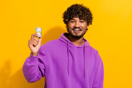 Portrait of a smiling young man in a purple hoodie holding rolled money against a yellow backgroundの写真素材