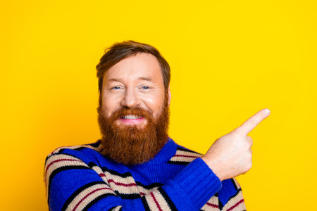 Smiling red-haired man with beard pointing right against yellow background wearing striped sweater.の写真素材